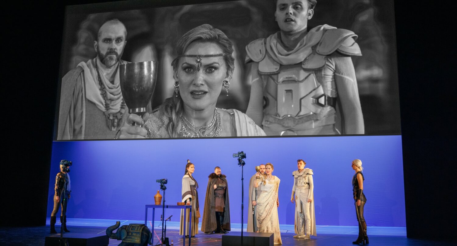 Thomas Walker, Mari Eriksmoen und Alexander Chance in: „Tamerlano“ von Georg Friedrich Händel am Badischen Staatstheater Karlsruhe. Auf eine große Leinwand oben wird das Geschehen auf der Bühne in schwarz-weiß projiziert.