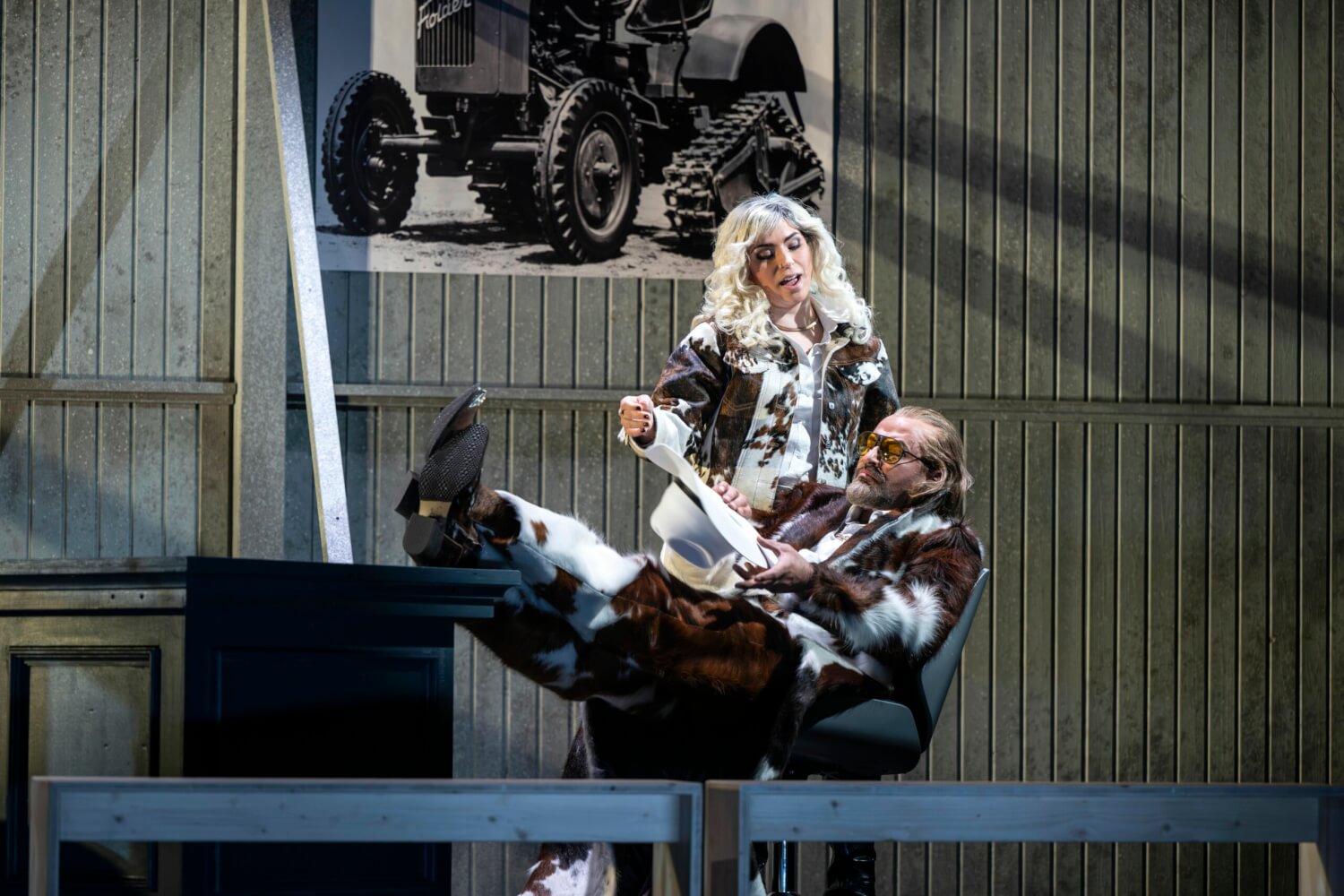 Aufführungsfoto von „Brokeback Mountain“ von Charles Wuorinen am Theater Erfurt. Ein Mann lehnt in seinem Schreibtischstuhl, die Füße auf dem Tisch abgelegt und hält Papiere in der Hand. Eine Fraz steht daneben.