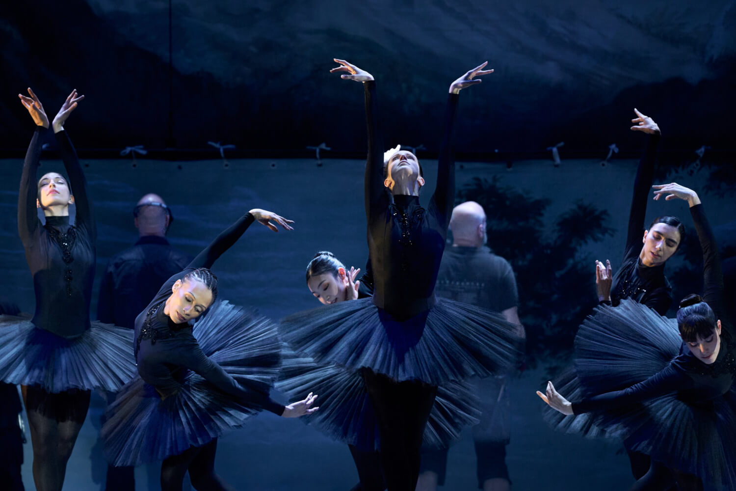 Aufführungsfoto von „Nurejew“ Staatsballett Berlin. Tänzerinnen und Tänzer in schwarzen hautengen Anzügen und Tütüs spreizen die Arme.