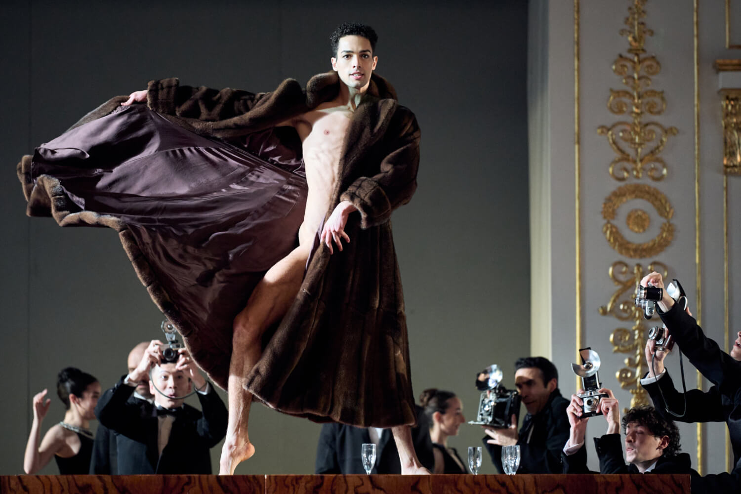 Aufführungsfoto von „Nurejew“ Staatsballett Berlin. Ein Mann hebt einen schweren, langen, dunkelbraunen Pelzmantel, unter dem er nackt ist, an. Drumherum Menschen mit alten Fotoapparaten.