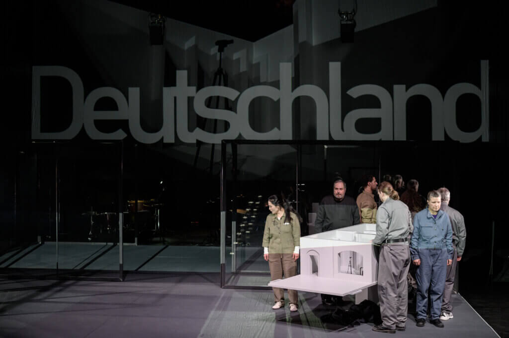 Im Hintergrund steht an der Wand in großen Buchstaben "Deutschland". Davor steht das Ensemble auf der rechten Seite an einer Art Tisch.