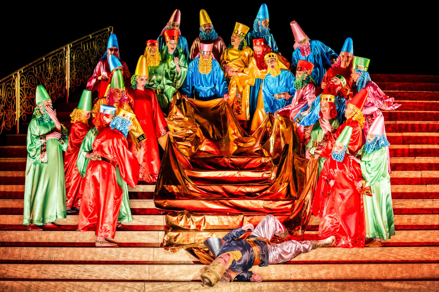 Aufführungsfoto von „Belshazzar“ von Georg Friedrich Händel, in der Regie von Herbert Fritsch an der Komischen Oper Berlin. Eine sehr bunt kostümierte Menschenschar steht auf einer großen Treppe und umringt einen Mann mit langer Schleppe, der auf den Stufen liegt.