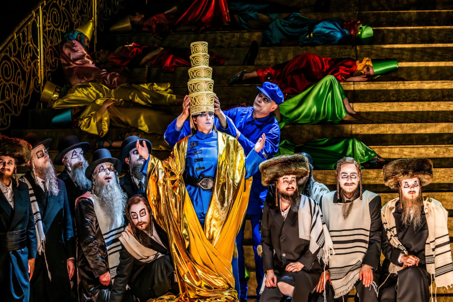 Aufführungsfoto von „Belshazzar“ von Georg Friedrich Händel, in der Regie von Herbert Fritsch an der Komischen Oper Berlin. Ein Mann im blauen Overall setzt einem anderen Mann mit goldenem Umhang eine riesige tortenähnliche Krone auf. Ein Chor aus weiß-gesichtigen Männern steht drumherum.