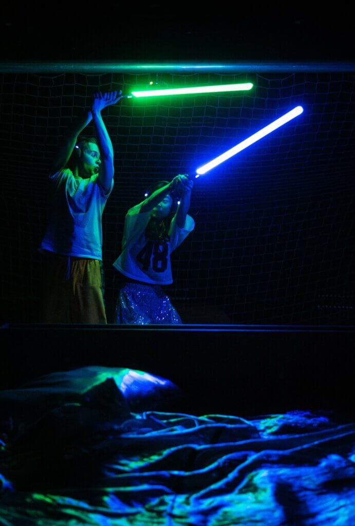 Aufführungsfoto von „Hall of Fans“ am Jungen Nationaltheater Mannheim, von Henrike Iglesias & Hempel mit Theo Teichmann und Soyi Cho. Im Dunkeln halten ein junger Mann und eine junge Frau zwei grün und blau leuchtende Laserschwerte in die Höhe.