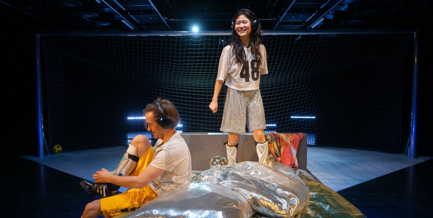 Aufführungsfoto von „Hall of Fans“ am Jungen Nationaltheater Mannheim, von Henrike Iglesias & Hempel mit Theo Teichmann und Soyi Cho. Auf einem Bett steht eine junge Frau mit Kopfhörern, auf der Bettkante sitzt ein junger Mann mit Kopfhörern und bindet sich die Fußballschuhe.