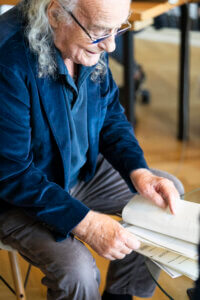 Roberto Ciulli in seinem Arbeitszimmer. Er sitzt auf einem Stuhl und blättert lächelnd in einer Blattsammlung.