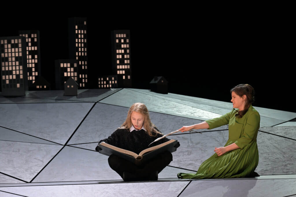 Iurii Iushkevich (The Boy) und Elizabeth Reiter (Agnès) sitzen alleine auf der Bühne. Iushkevich im Schneidersitz mit einem aufgeschlagenen Buch auf dem Schoß, Reiter daneben, ihm zugewandt.