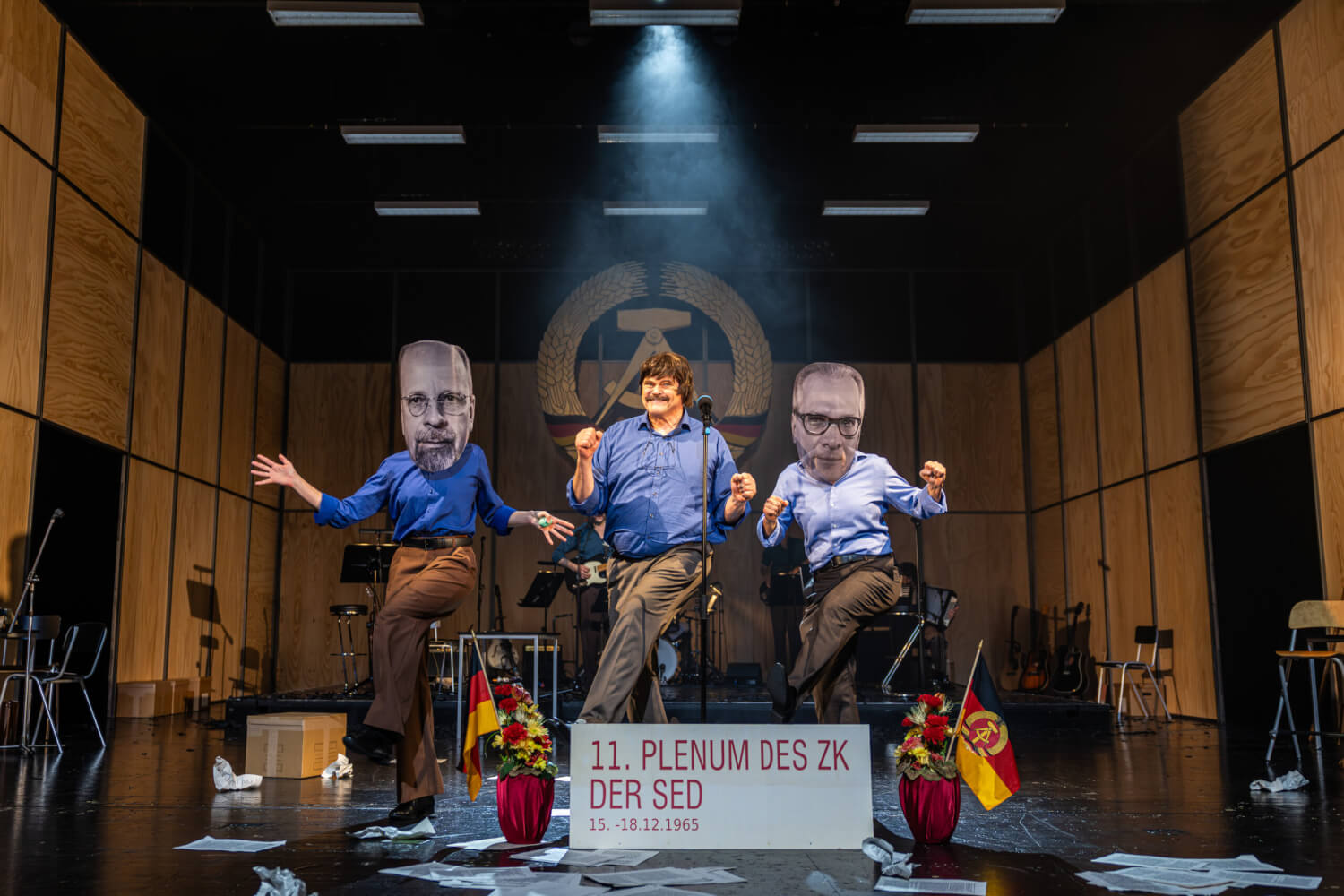 Aufführungsfoto von „Biermann – Drachentöter“ von Martin Heckmanns am Staatstheater Meiningen. Im Hintergrund das Emblem der DDR. Im Vordergrund ein Mann, der aussieht wie Wolf Biermann. Neben ihm tanzend, zwei Personen, die riesige Pappmasken-Gesichter von Erich Honecker und Walter Ulbricht tragen.