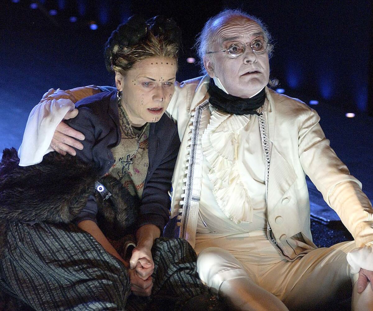 Schauspieler Roberto Ciulli in „Die Wände“. Ein älterer Mann im weißen Anzug blickt mit aufgerissenen Augen nach vorne und hält eine Frau, die rechts neben ihm sitzt im Arm.