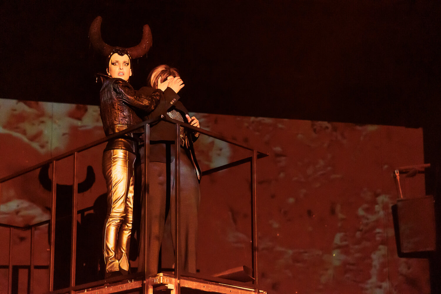 Mephisto steht links mit Dr. Margarete Faust an einer Brüstung und hält ihr die Augen zu.