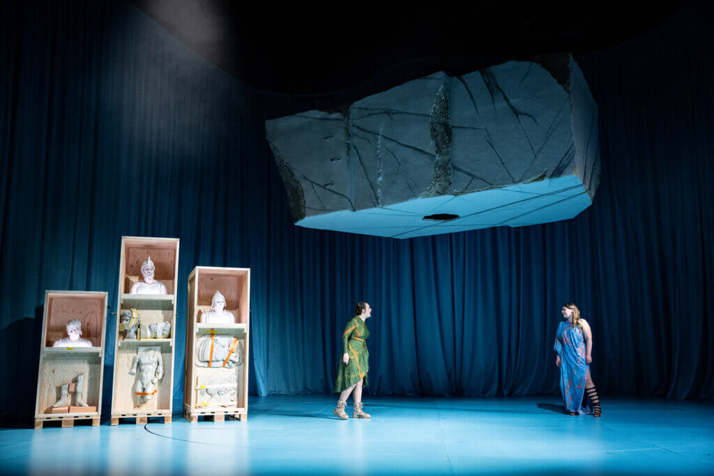 Auf der Bühne stehen links drei Regale. Rechts schauen sich zwei Spieler an, während über ihnen ein riesiger Felsbrocken bedrohlich schwebt.