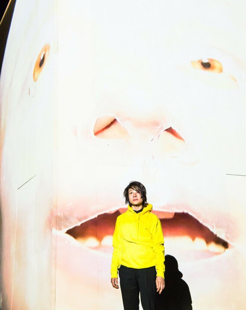 „Fanny und Alexander“ nach dem Film von Ingmar Bergman am Düsseldorfer Schauspielhaus. Eine junge Person mit knallgelber Jacke steht vor einer riesigen Videoleinwand, auf der ein verzerrtes Gesicht zu sehen ist.