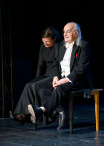 Aufführungsfoto von „Gespenster“ mit Roberto Ciulli. Ein alter Mann im Frack sitzt auf einem Tisch mit baumelnden Beinen. Neben ihm eine Frau im schwarzen Kleid.