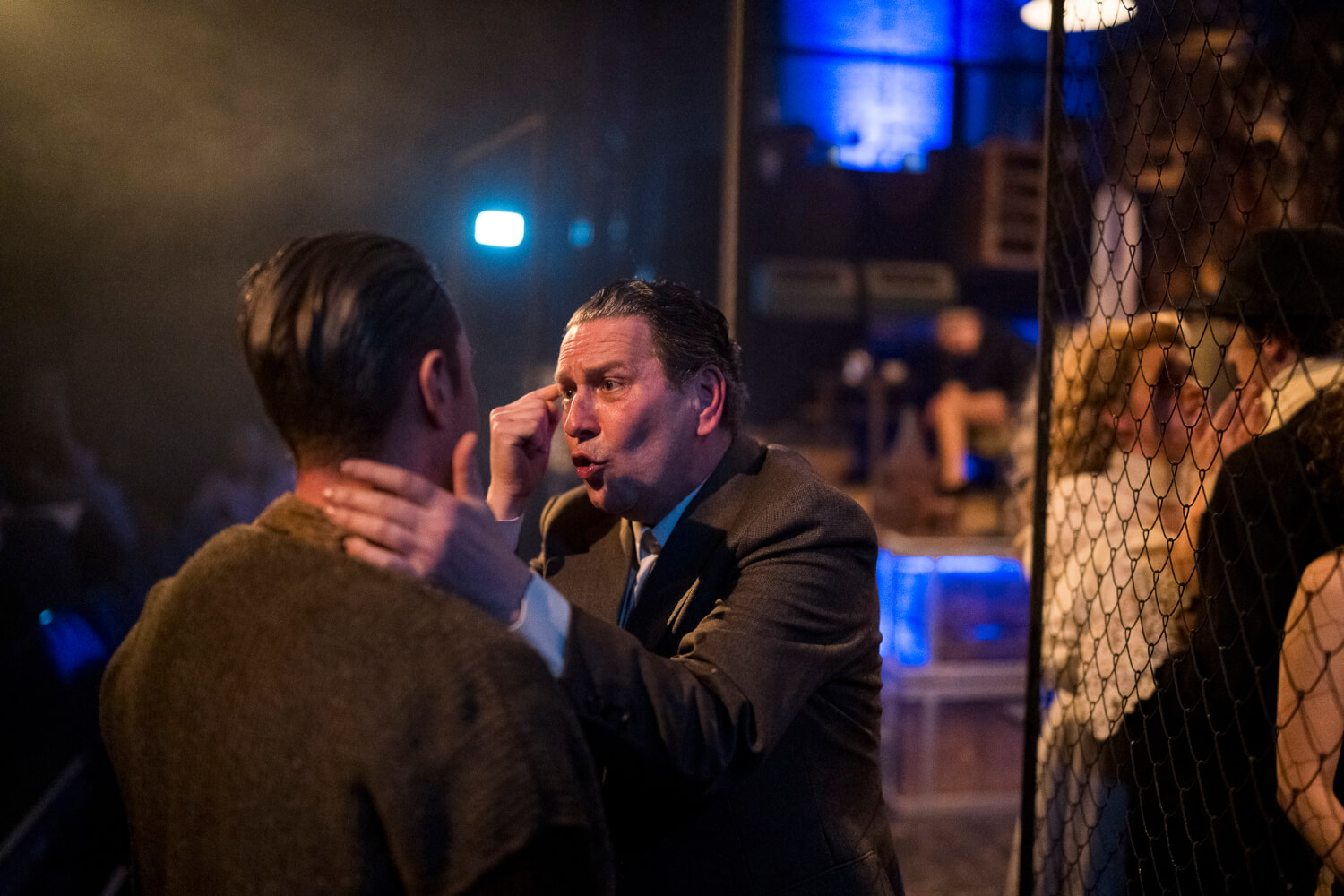 Aufführungsfoto von „Von dem Glück, Hrdlak gekannt zu haben“ am Gerhart Hauptmann-Theater Göritz-Zittau. Ein Mann tippt sich mit dem Zeigefinger an die Schläfe und umfasst mit der anderen Hand einen Mann am Nacken, dabei sieht er wütend-verzweifelt aus.