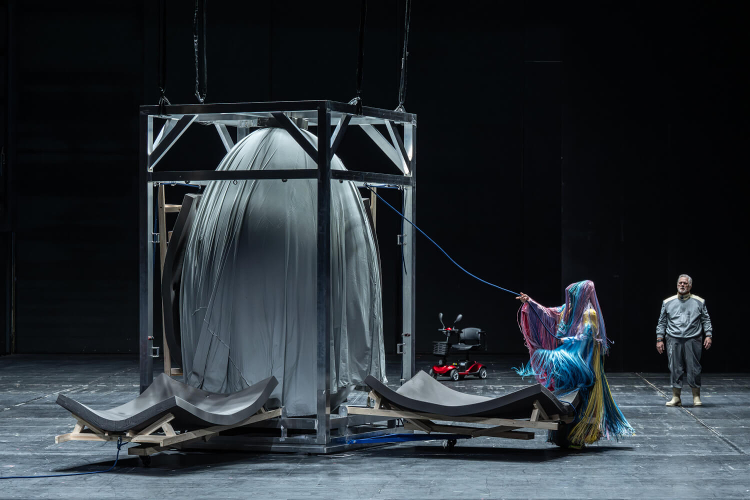 „Die große Stille“ von Christopher Rüping nach Wolfgang Amadeus Mozart an der Staatsoper Hamburg mit Ana Durlovski und Gregory Kunde. In einem Metallgestell steht ein mit Stoff umhüllter Gegenstand, der wie ein Ei aussieht. Eine mit bunten Stoffen behangene Frau sieht an einem Seil, das mit dem Gestell verbunden ist.