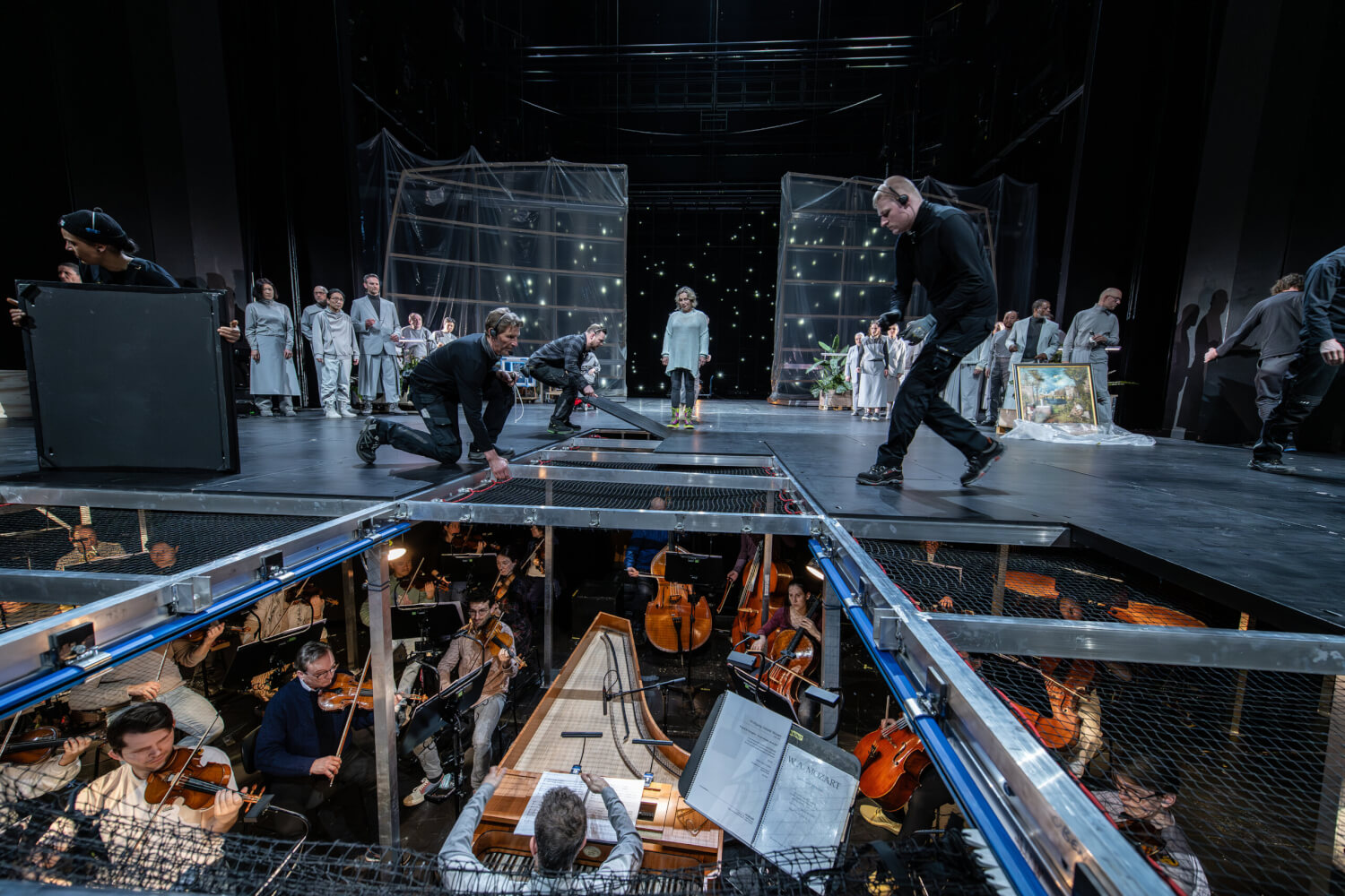 „Die große Stille“ von Christopher Rüping nach Wolfgang Amadeus Mozart an der Staatsoper Hamburg. Bühnentechniker heben Bühnenbodenplatten weg. Darunter sieht man das Orchester spielen. Im Hintergrund auf der Bühne eine Sängerin in der Mitte und recht und links der Chor.