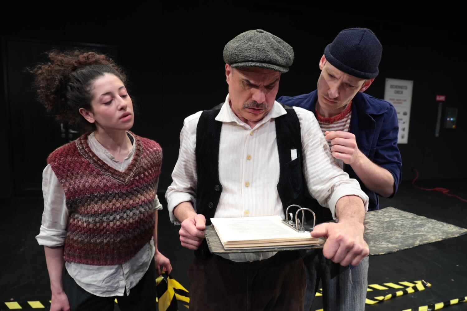 Aufführungsfoto von „Eintritt auf eigene Gefahr“, Stückentwicklung von Grete Pagan am Jungen Ensemble Stuttgart. Ein Mann mit schwarzer Weste, weißem Hemd und Mütze schaut in einen Aktenordner, ein Mann und eine Fraz stehen daneben und gucken ihm über die Schulter.