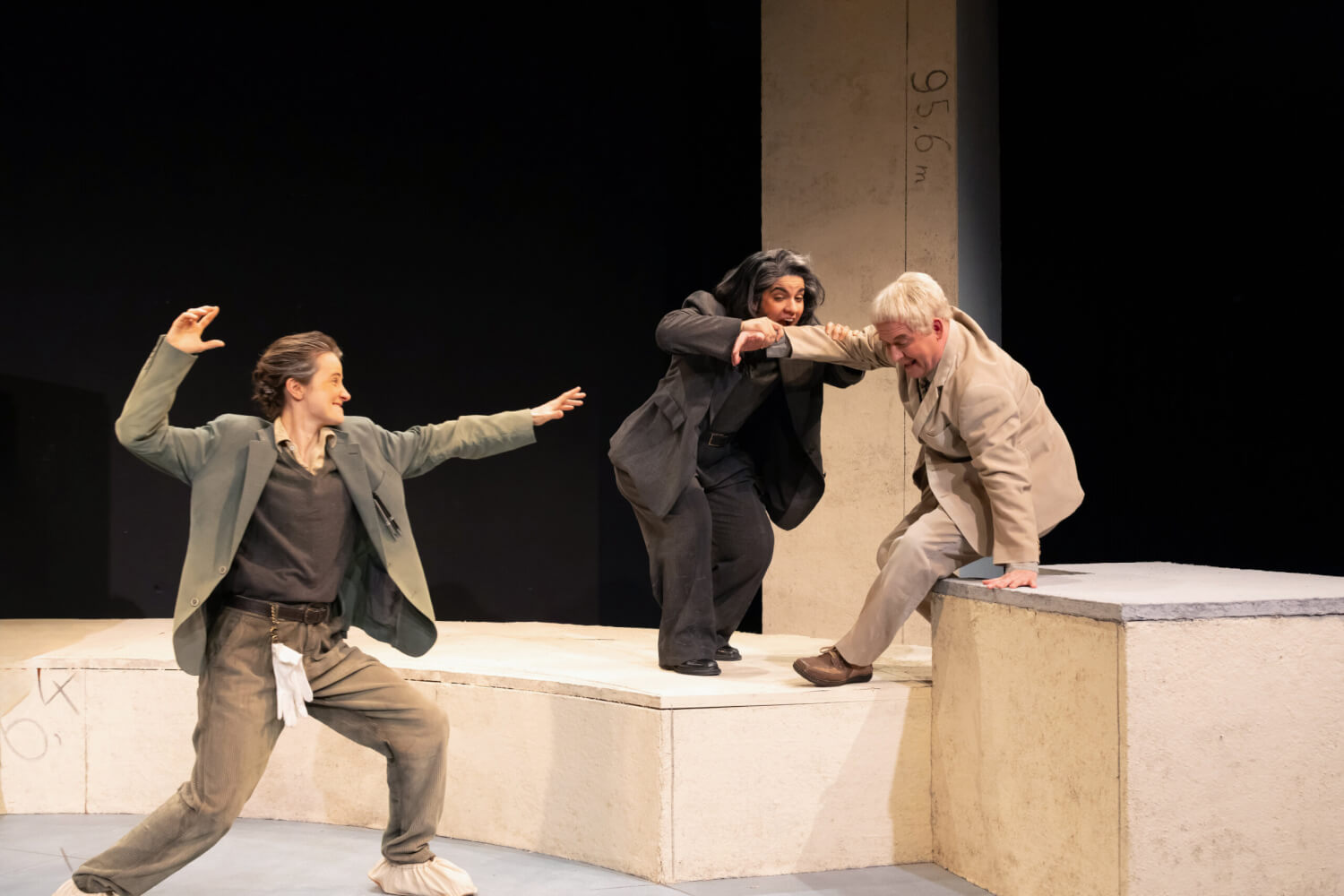 Aufführungsfoto von „Ehrlich“ von Jörn Klare am Anhaltischen Theater Dessau. Eine Frau holt mit ihren Armen zum Schlag aus, eine andere Frau beißt dem Mann in seinen rechten Arm, beide stehen auf Holzkästen.