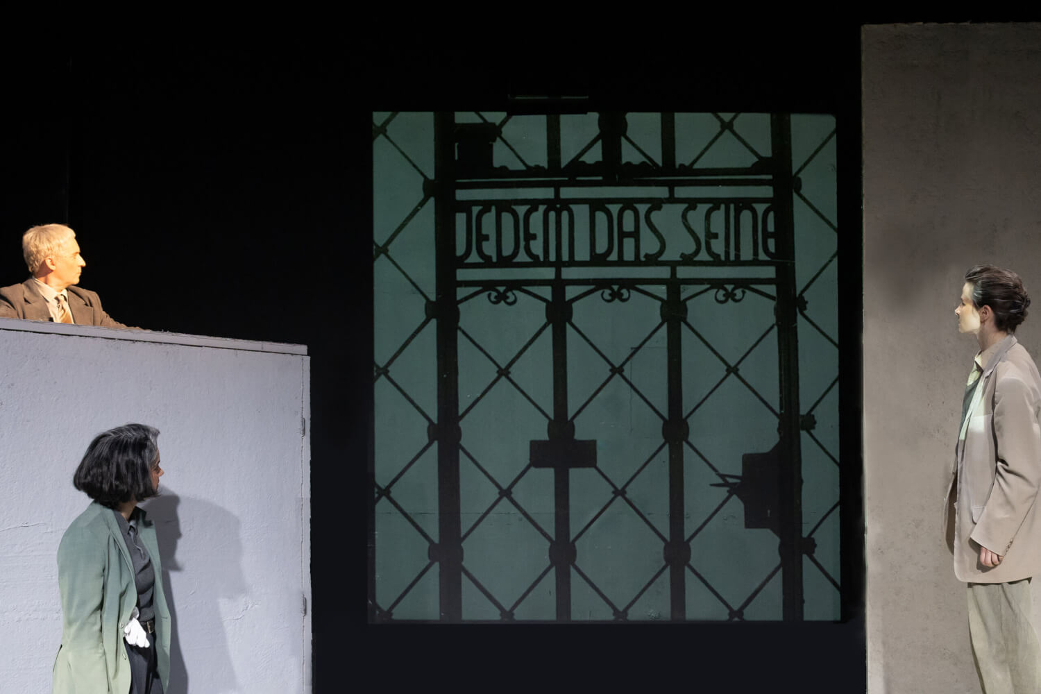 Aufführungsfoto von „Ehrlich“ von Jörn Klare am Anhaltischen Theater Dessau. In der Mitte der Schattenriss eines Tores mit der Aufschrift Jedem das seine. Zwei Frauen und ein Mann blicken den Schattenriss in der Mitte an.