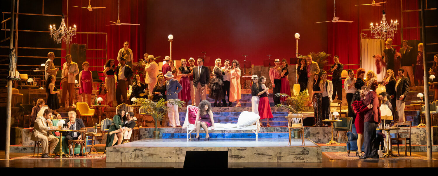 Die Bühne ist prall gefüllt mit Darstellenden und Requisiten, fast wie bei einem Wimmelbild, wo man Gegenstände suchen muss. In der Mitte im Scheinwerferlicht sitzt Alma auf einer Chaiselongue.