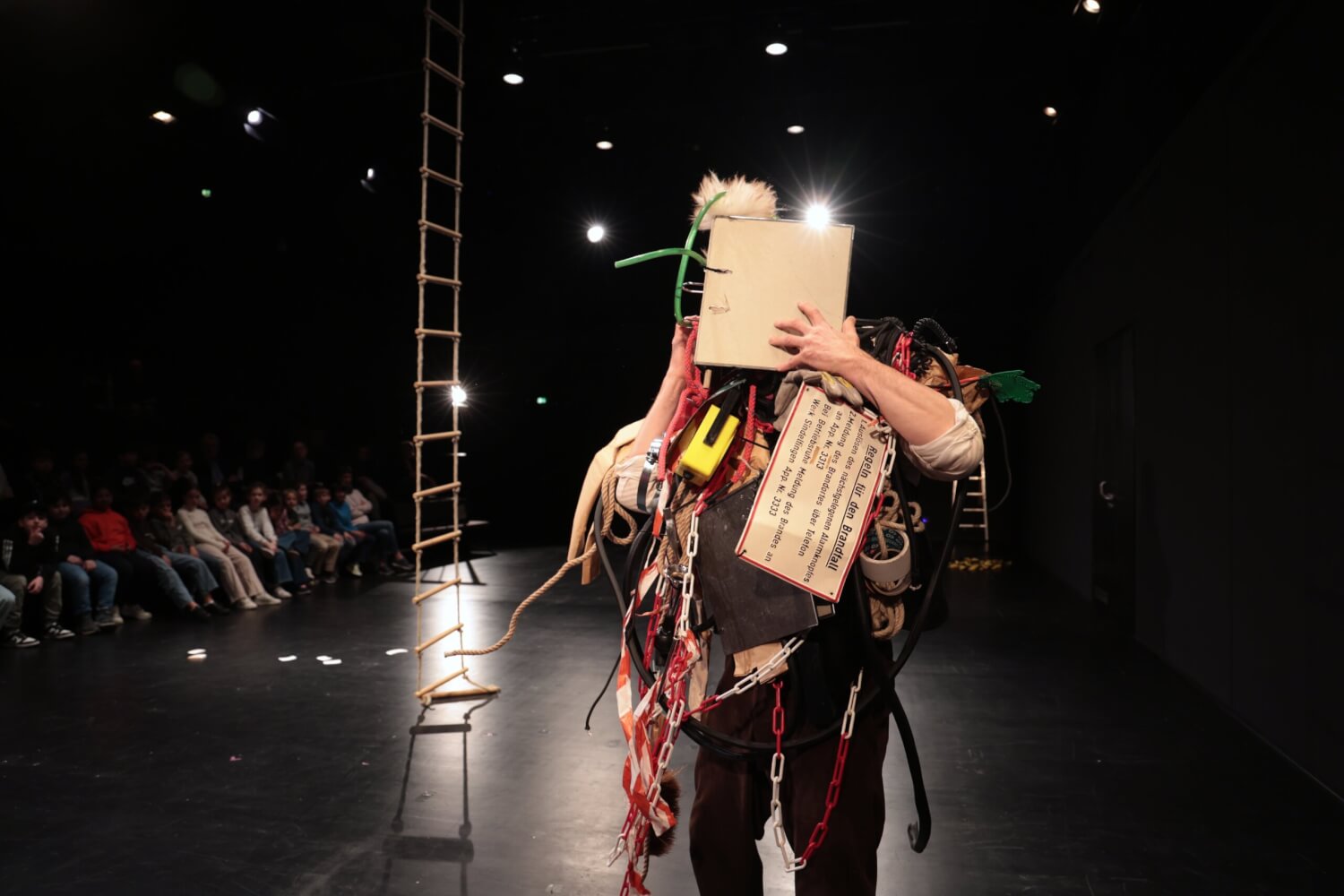 Aufführungsfoto von „Eintritt auf eigene Gefahr“, Stückentwicklung von Grete Pagan am Jungen Ensemble Stuttgart. Ein Mann ist von oben bis unten bepackt und behägt mit Absperrband, Absperrkette, Seil, einem Schild mit Regeln im Brandfall und einem Ordner vor dem Gesicht.