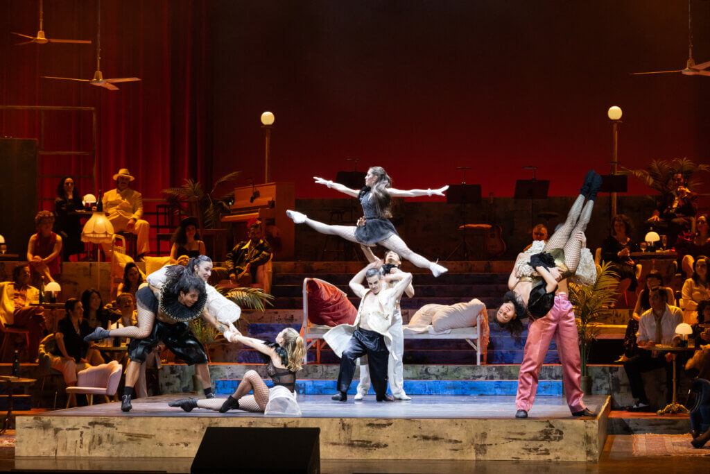 Das Ballettensemble steht auf einer rot ausgeleuchteten Bühne und zeigt unterschiedliche Hebefiguren.