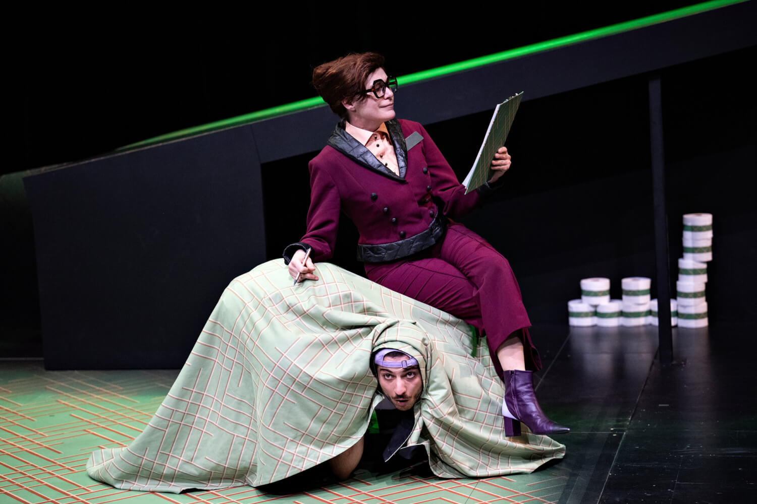 Aufführungsfoto von „Hotel Lauter und die Unsichtbaren“ von Armela Madreiter am Theater Heidelberg. Eine Frau im bordeauxrotem Anzug sitzt mit einem Klemmbrett und Stift in der Hand auf einem Stoffhaufen, unter dem der Kopf eines jungen Mannes hervorschaut.