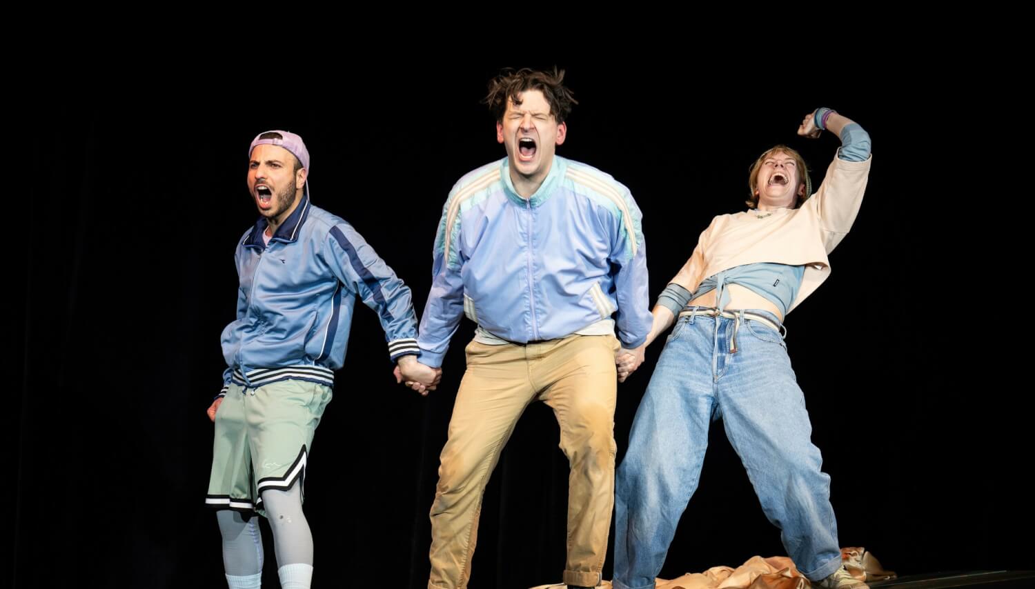 Aufführungsfoto von „Hotel Lauter und die Unsichtbaren“ von Armela Madreiter am Theater Heidelberg. Zwei junge Männer und eine junge Frau halten sich an den Händen nebeneinander auf einer leichten Schrägen und schreien und lachen.