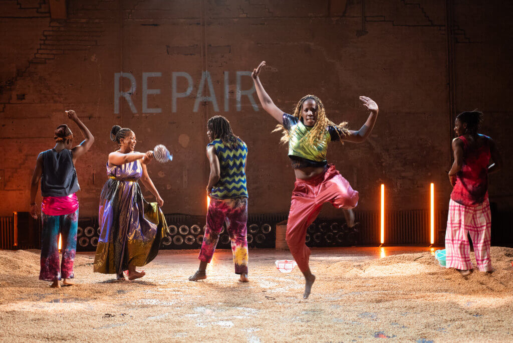 Fünf Tänzer:innen stehen in einer Reihe nebeneinander. Die zweite Person von rechts springt mit freudig erhobenben Armen vom Boden hoch.