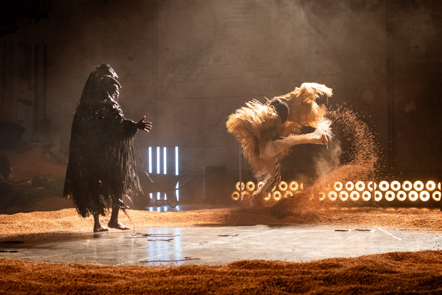 Links steht eine dunkel vermumte Person in weiter Kleidung und wendet sich einer weiteren Tänzer:in zu, die Sand aufwirbelt.