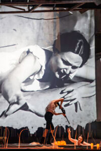 Schwarzweißbild einer weiblichen Person mit ausgestreckter Hand. Davor auf der Bühne ein Darsteller mit Waffe, der eine weiter Darstellerin bedroht