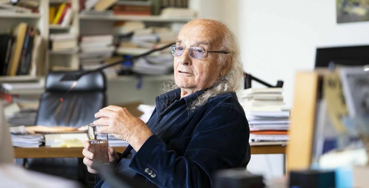 Roberto Ciulli sitzt in seinem Arbeitszimmer vor einer unordentlichen Bücherwand, mit einem Glas Wasser in der Hand.