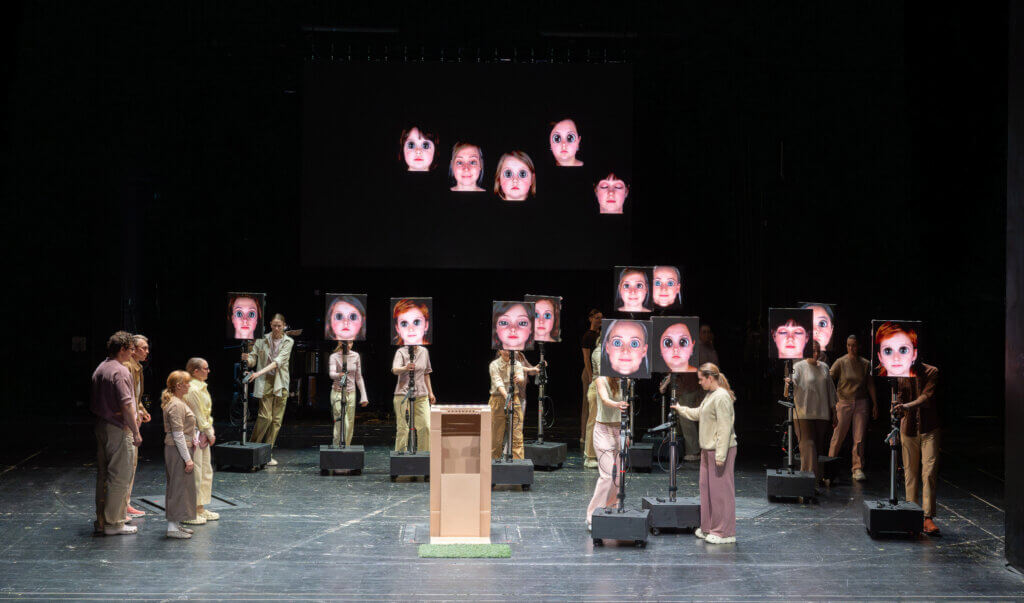 Auf der Bühne stehen unterschiedliche Expert:innen mit übergroßen Bildschirmen als Gesichter. Sie können zum Sprechen nach vorne gezogen werden. In der Mitte steht vorne ein Redner:innenpult.