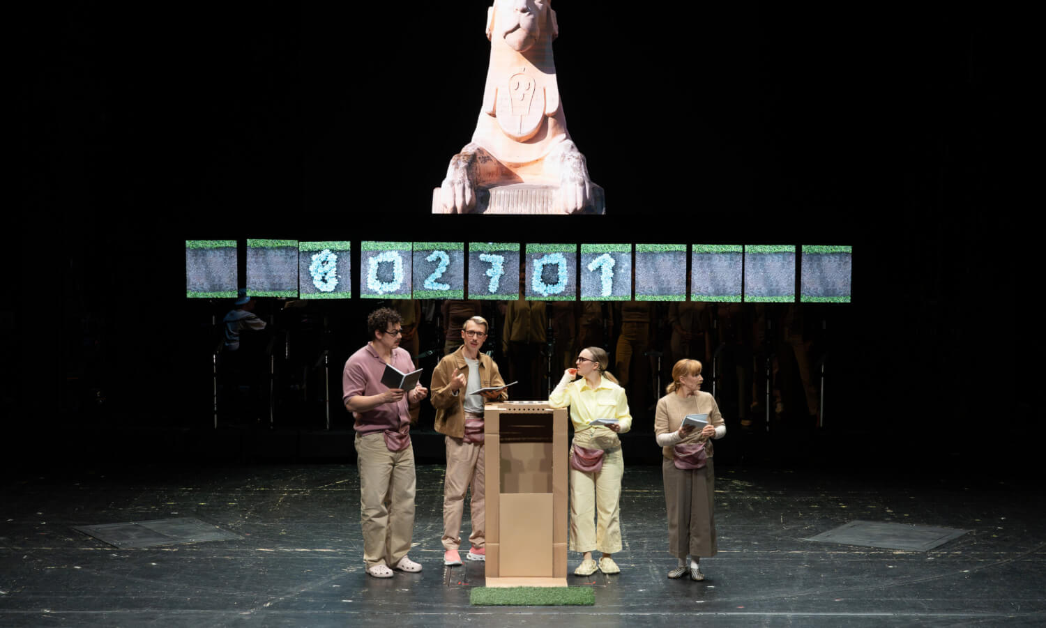 Das Ensemble steht in der Mitte der Bühne hinter einem Redner:innenpult. Im Hintergrund hängt ein Zeitstrahl.