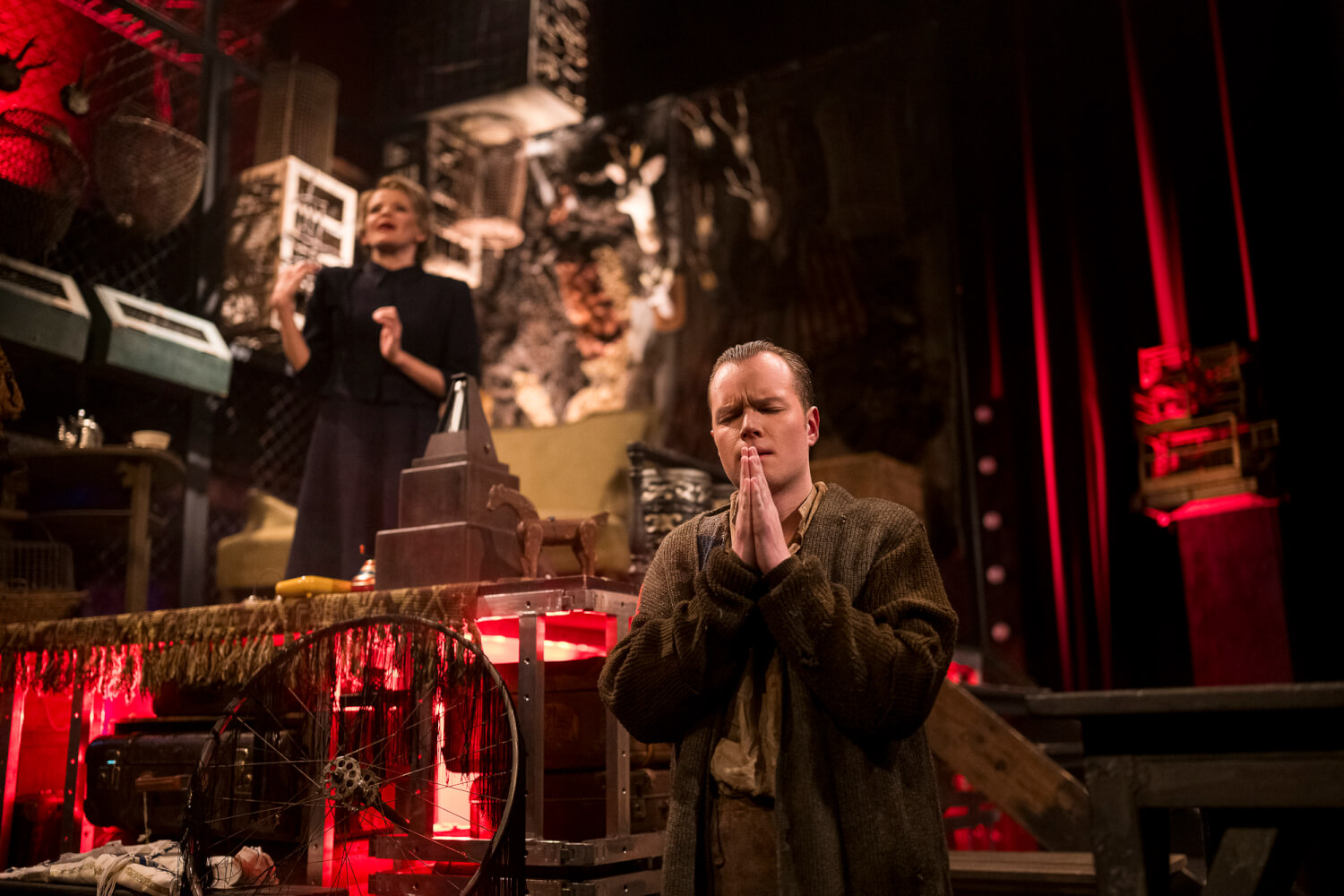 Aufführungsfoto von „Von dem Glück, Hrdlak gekannt zu haben“. Im Vordergrund steht ein Mann mit geschlossenen Augen und zum Gebet gefalteten Händen. Im Hintergrund sitzt eine Frau erhöhrt und blickt nach vorne.