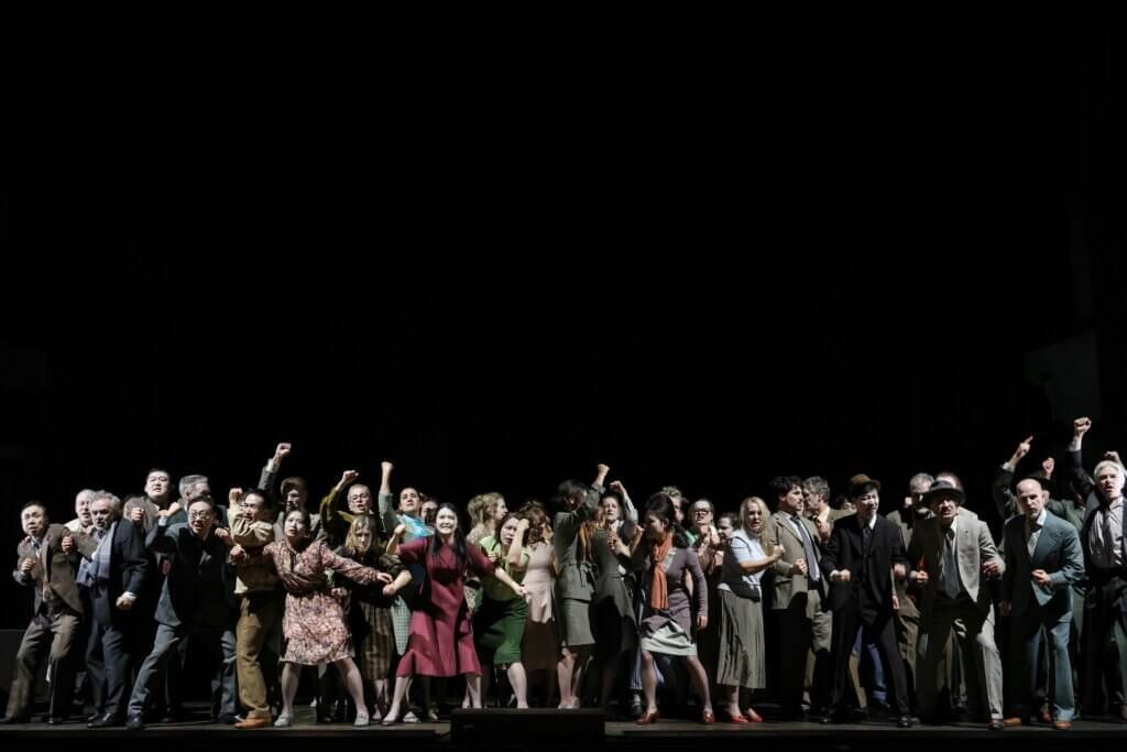 Der Chor steht in einer bunten Menge auf der Bühne, Gesichter nach oben gewandt.