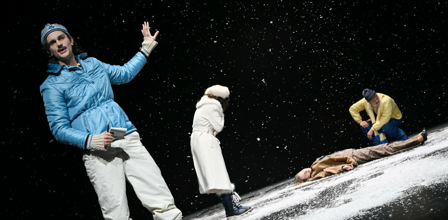 Vorne links steht eine Person in Winterkleidung. Auf der Bühne liegt Schnee. Hinten rechts liegt jemand regungslos auf dem Boden, zwei Personen beugen sich über den Körper.