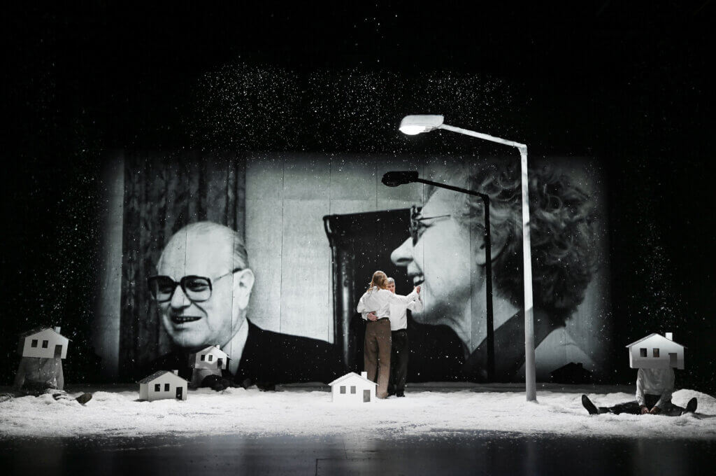 Das Ensemble ist auf einer verschneiten Bühne verteilt. Manche sitzen mit weißen Häuser-Masken über den Köpfen auf dem Boden. Im Hintergrund wird ein Bild von Shlomo Lewin und Frida Poeschke angestrahlt.