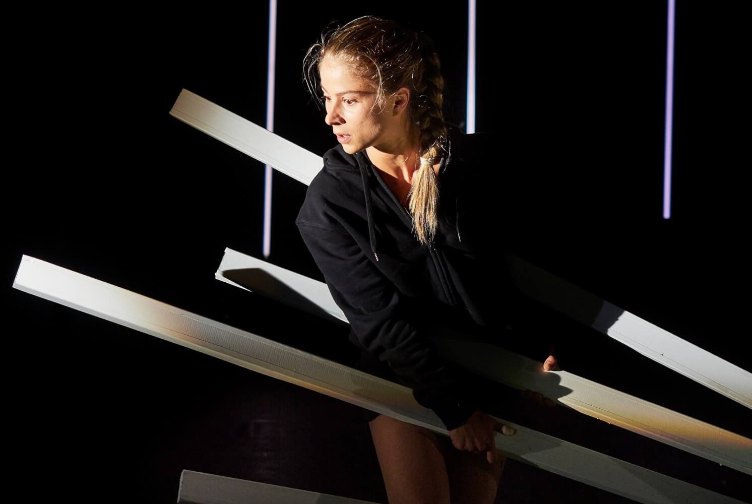 Aufführungsfoto von „Bungalow“ nach Helene Hegemann am Düsseldorfer Schauspielhaus mit Lea Ruckpaul als Charlie. Auf dunkler Bühne sitzt eine junge Frau zwischen hell angeleuchteten Holzbalken.