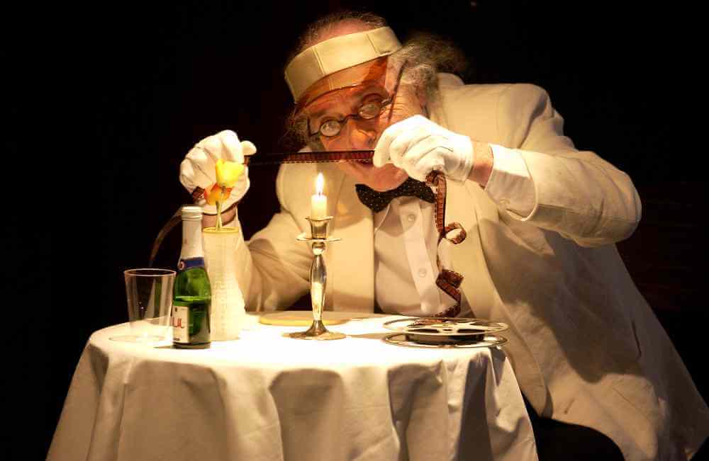 Aufführungsfoto von „Es geht immer besser, besser – immer besser…“ mit Roberto Ciulli. Ein alter Herr mit weißem Anzug sitzt an einem kleinen runden Tisch mit weißer Tischdecke, auf dem eine Kerze und eine kleine Flasche Sekt stehen. Er hält einen Foto-Negativstreifen über die brennende Kerze.