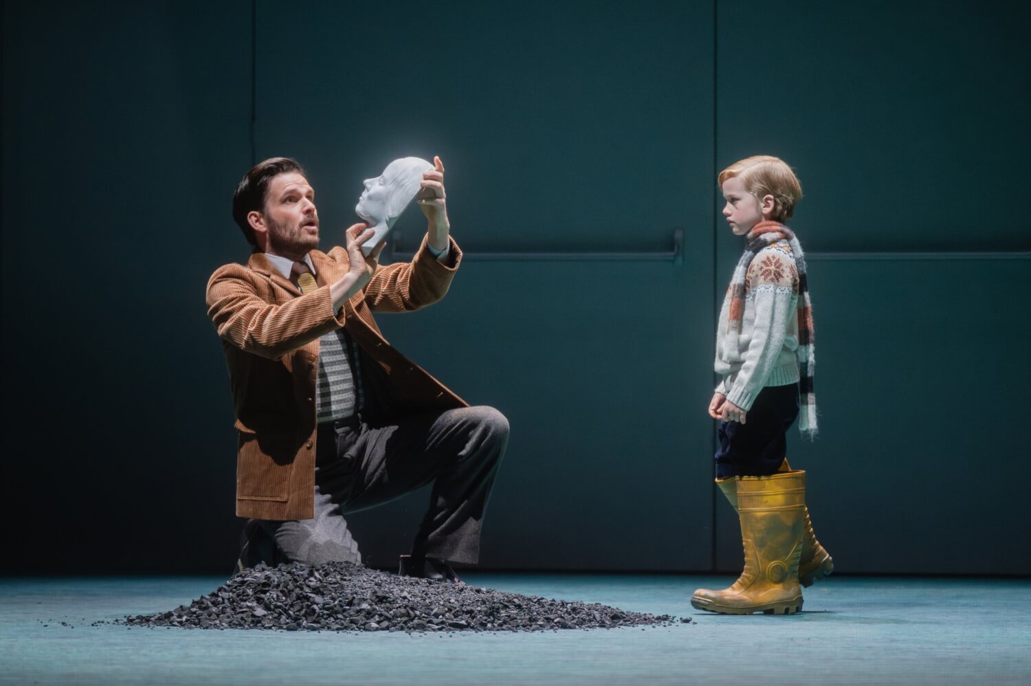 Aufführungsfoto von „Coming Up for Air“ von Bernd Franke an der Oper Leipzig. Ein Mann kniet mit einer Gipsmaske in den Händen vor einem kleinen Jungen in zu großen Gummistiefeln, der ihn anschaut.