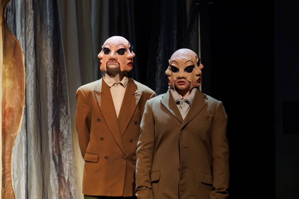 Zwei Männer stehen mit Gesichtsmasken und Anzug nebeneinander vor einem Vorhang. Die Masken erzeugen die Illusion, die Personen hätten jeweils drei Gesichter: eins nach vorn, eins nach links und eins nach rechts.