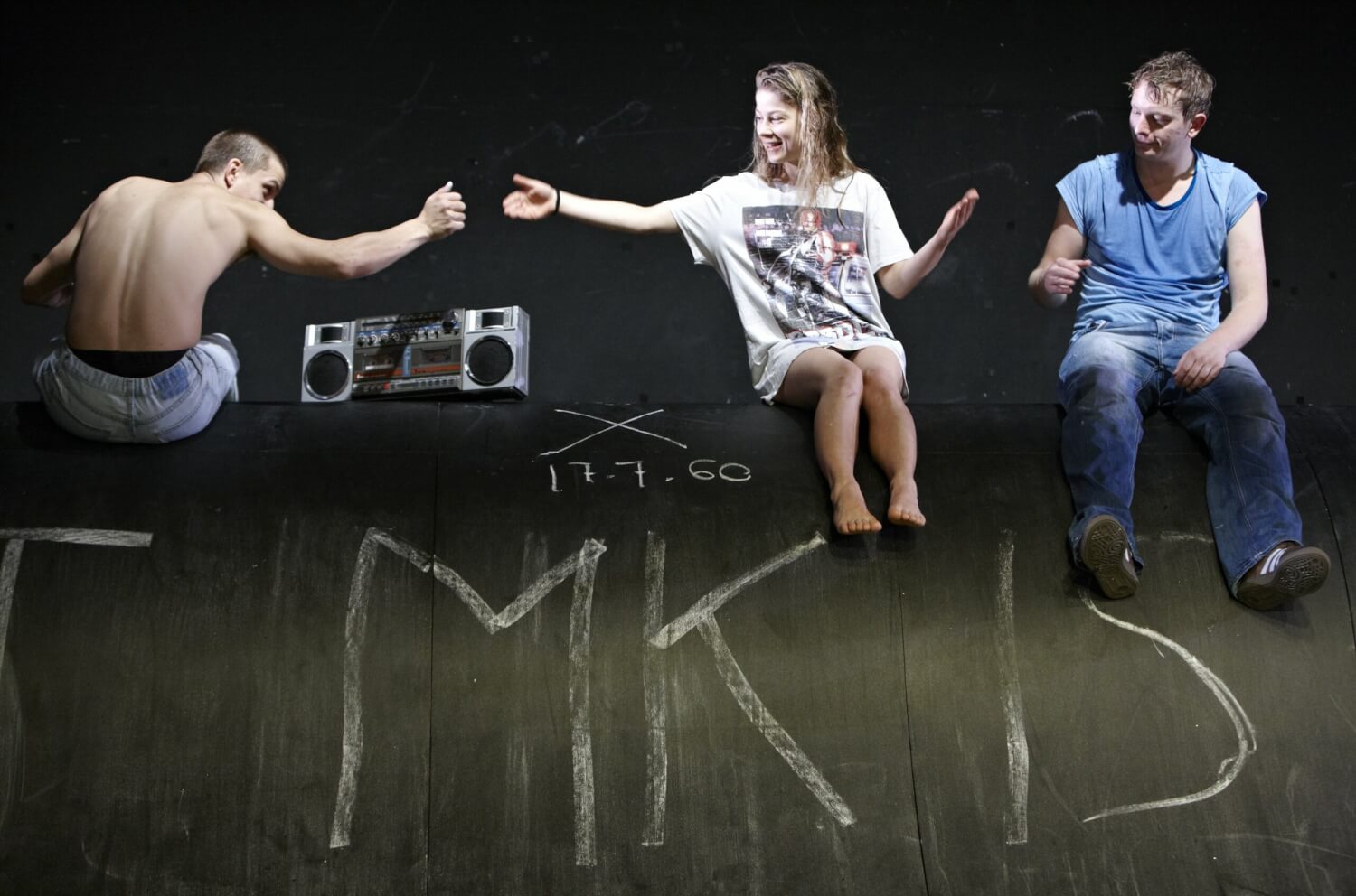 Aufführungsfoto von „Tschick“ von Wolfgang Herrendorf am Staatsschauspiel Dresden. Auf einer dunklen Schieferrampe sitzen ein junger Mann mit dem Rücken zum Betrachter, eine junge Frau, die ihm die rechte Hand entgegenstreckt und lächelt und recht ein anderer junger Mann.
