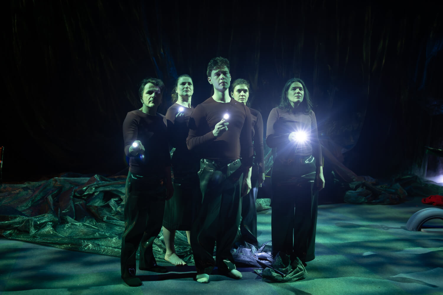 Das Ensemble steht aneinander gedrängt in der Mitte der Bühne und leuchtet mit Taschenlampen nach vorn.