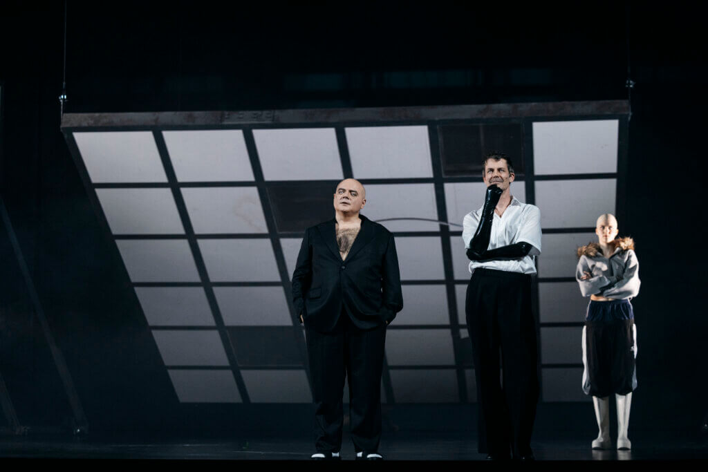 Drei Darstellende stehen vor einer schrägen Ebene aus Fensterglas. Alle tragen weiße Kontaktlinsen.