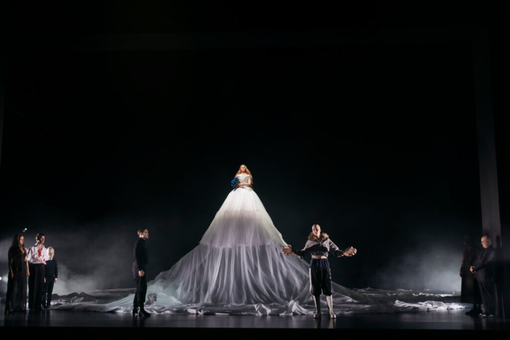 Inga Schaefer steht auf einer erhöhten Ebene in einem gigantischen Brautkleid. Es reicht über die Ebene bis auf den Boden. Rechts und links vor ihr stehen zwei weitere Darstellende.