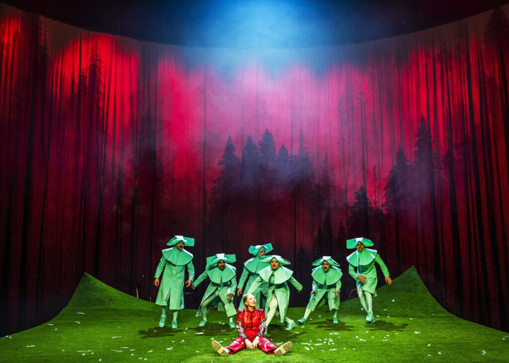Eine Person sitzt in roter Kleidung auf dem Boden, die Beine von sich gestreckt. Dahinter steht ein Ensemble in grünen Mänteln. Die Lichtung ist in rotes, Licht getaucht und ragt bedrohlich hinter dem Ensemble auf.