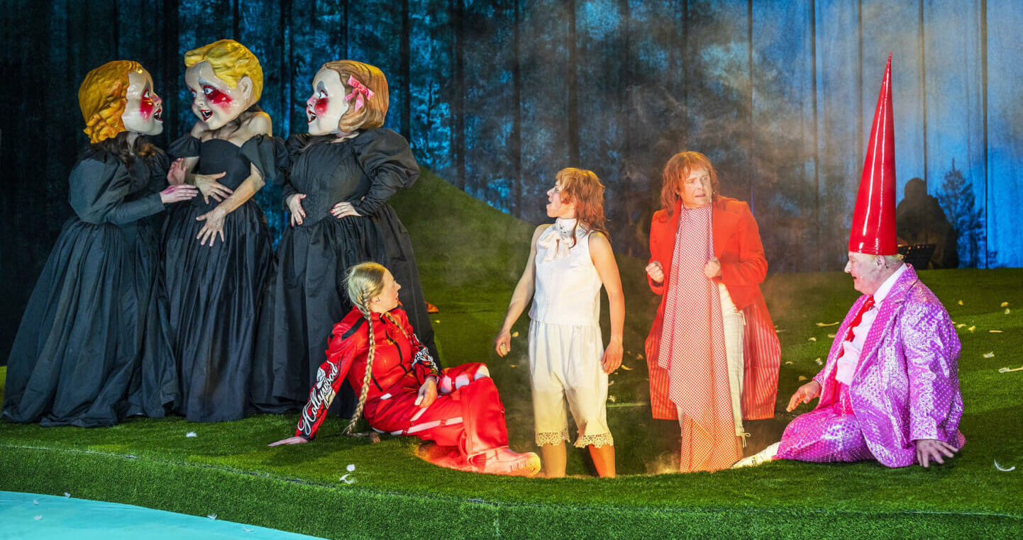 Die Gebrüder Grimm stehen inmitten einer bunten Truppe und schauen staunend nach links und rechts. Um sie herum ist eine nebelige Waldlichtung angelegt. Links stehen drei Personen in langen Kleidern mit überdimensionalen Köpfen und unterhalten sich.