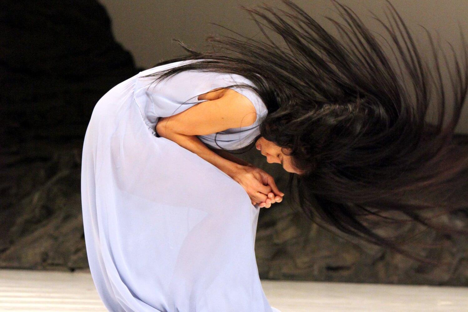 Ditta Miranda Jasjfi in Pina Bauschs Stück „Masurca Fogo“. Das Bild wurde 2012 in Wuppertal aufgenommen. Eine Frau mit langen schwarzen Haaren krümmt sich hockend, die Hände auf den Knien zusammengefaltet, den Kopf zu den Händen geneigt, die Haare wirft sie wir in einem Bogen vom Rücken nach vorne.