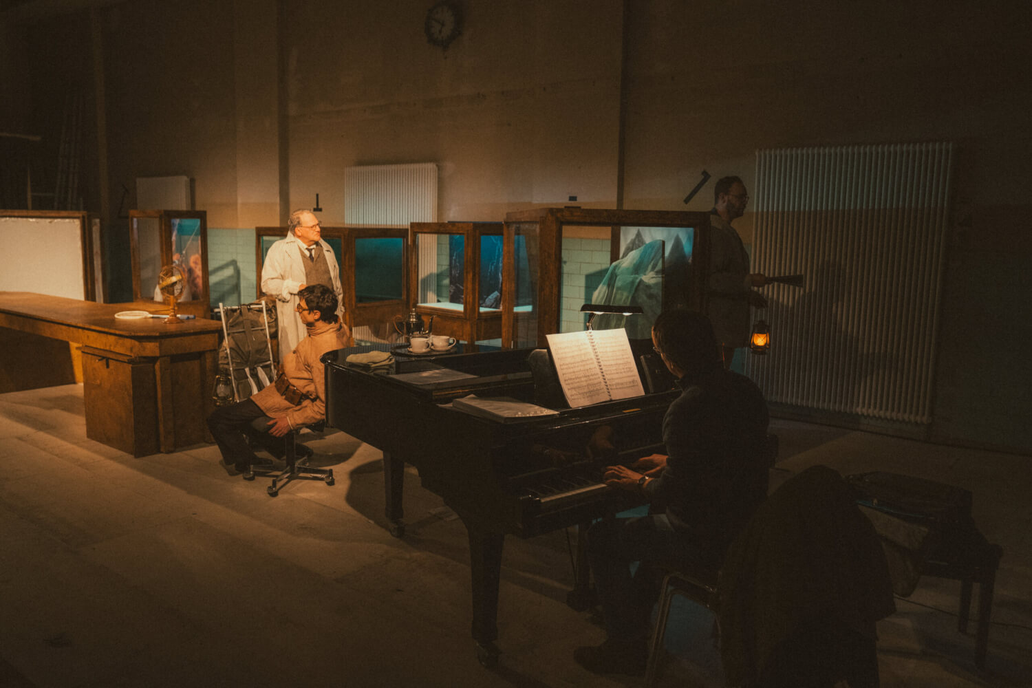 Probenfoto von „Die Reise zum Mittelpunkt der Erde“ am Staatstheater Augsburg. Ein Mann sitzt am Klavier. Ein Mann sitzt auf einem Schreibtischstuhl, ein anderer steht mit einem Arztkittel hinter ihm. An der Seite des Klaviers steht ein Mann mit Laterne in der Hand.